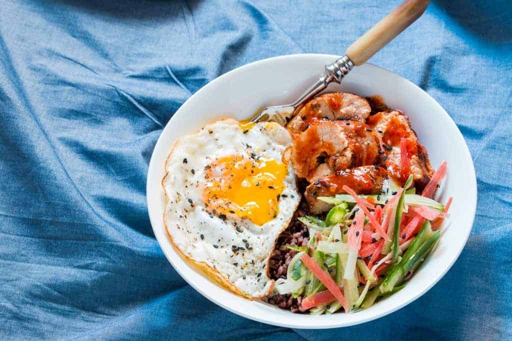 a white bowl of schezwan chicken, sliced veggies, black rice and fried egg