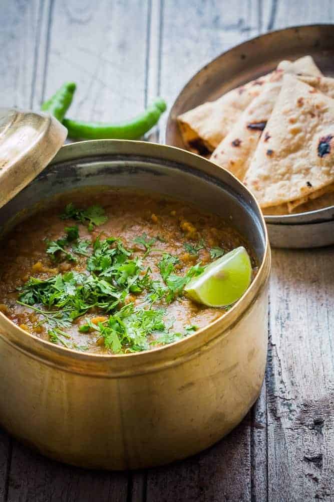 dhaba style dal in a brass container with a piece of lemon, garnished with coriander along with a plate of rotis and a few chillies on the side