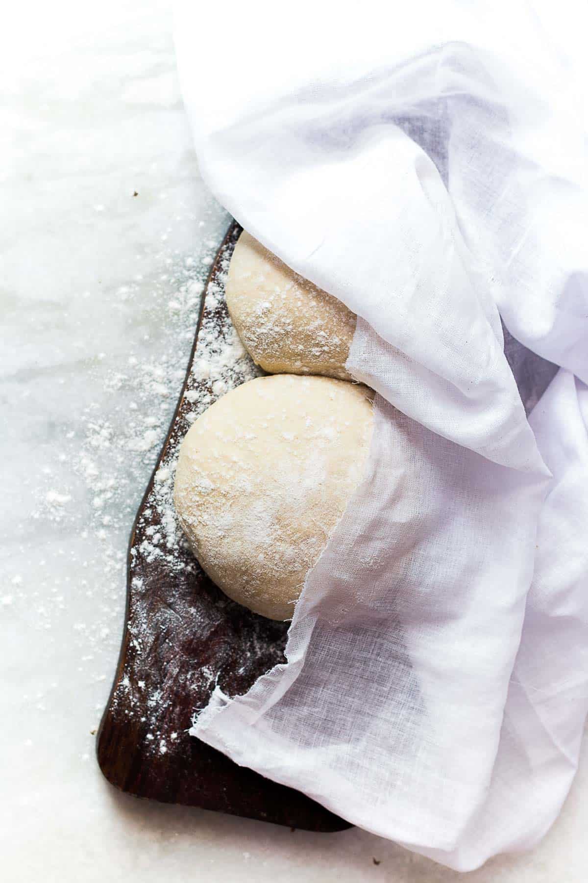 Pizza doughs placed on a dark brown board with a white cloth placed across it