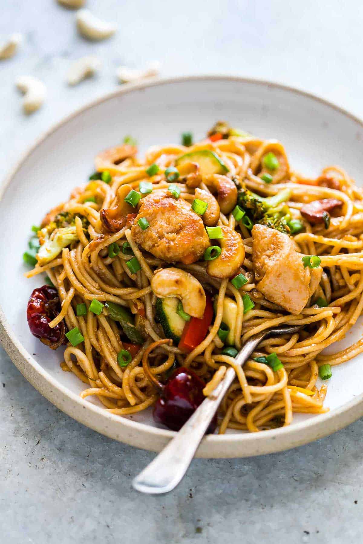 Chinese Cashew Chicken Noodles Stir-Fry garnished with sesame seeds, green onion and served in a white plate.