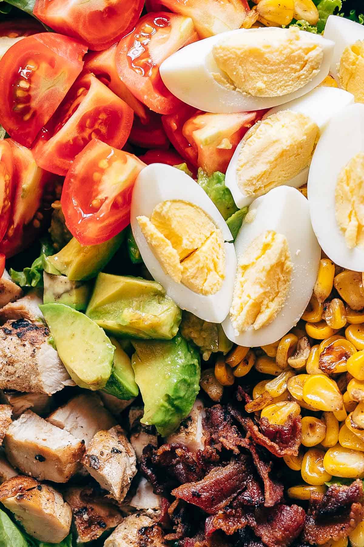 This is a classic layered chicken cobb salad recipe that has all the right fixings that includes lettuce, boiled eggs, grilled corn, bacon, grilled chicken, tomatoes and avocado! Serve this with a creamy avocado blue cheese dressing that's made with greek yogurt and you have dinner sorted. This isn't just a salad, it's a meal! Perfect for a crowd. Use steak instead of chicken or make it vegetarian #salad #recipe #chicken #dinner #easy Chicken Cobb Salad served in a bowl - it has lettuce, tomatoes, chicken, bacon, corn and boiled eggs