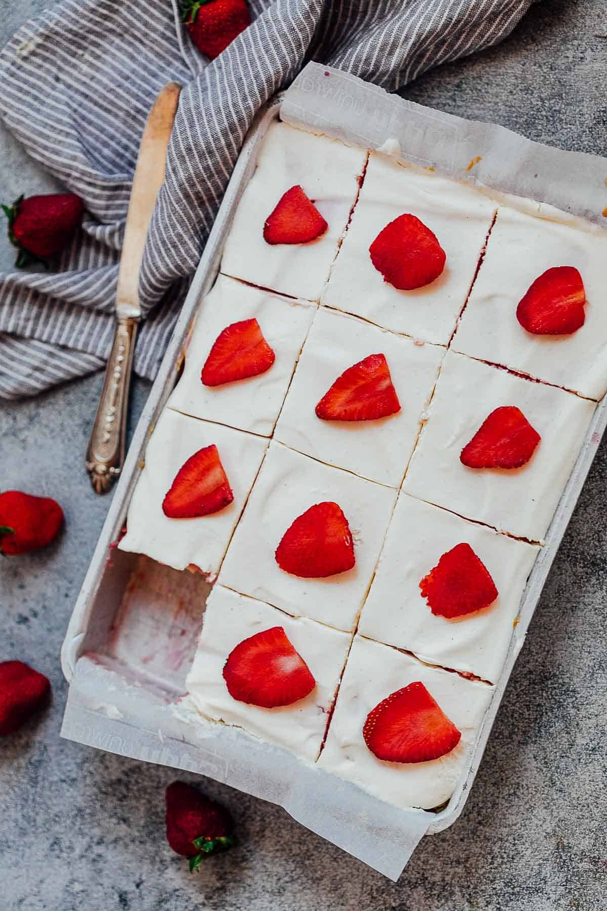 This strawberry poke cake is made from scratch and is a soft, moist jello poke cake that is topped with whipped cream and fresh strawberries. It has the taste of strawberries in every bite and is the perfect easy, elegant dessert! #dessert #cake #strawberry #easter #mothersday #pink Slices of strawberry poke cake in a sheet pan with a knife and fresh strawberries on the side