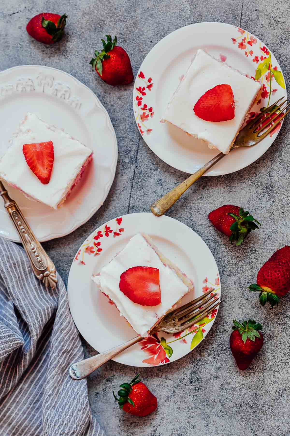 Three slices of strawberry poke cake made from scratch served on white plates with forks and fresh strawberries on the side
