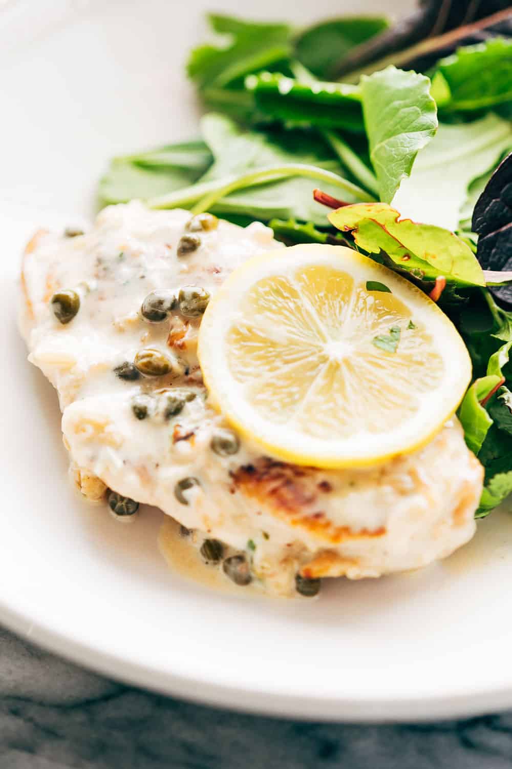 LEMON CHICKEN SCALLOPINI is a super quick, healthy and fast dinner recipe that takes less than 30 minutes to put on the table and has a delicious lemon sauce that you'll want to drink straight from the pan! Studded with capers, this dish looks fantastic. Its gluten free and keto friendly. #fastdinner #chicken #lunch #lemon #summer Creamy lemon chicken scallopini served with a light salad on the side in a white bowl