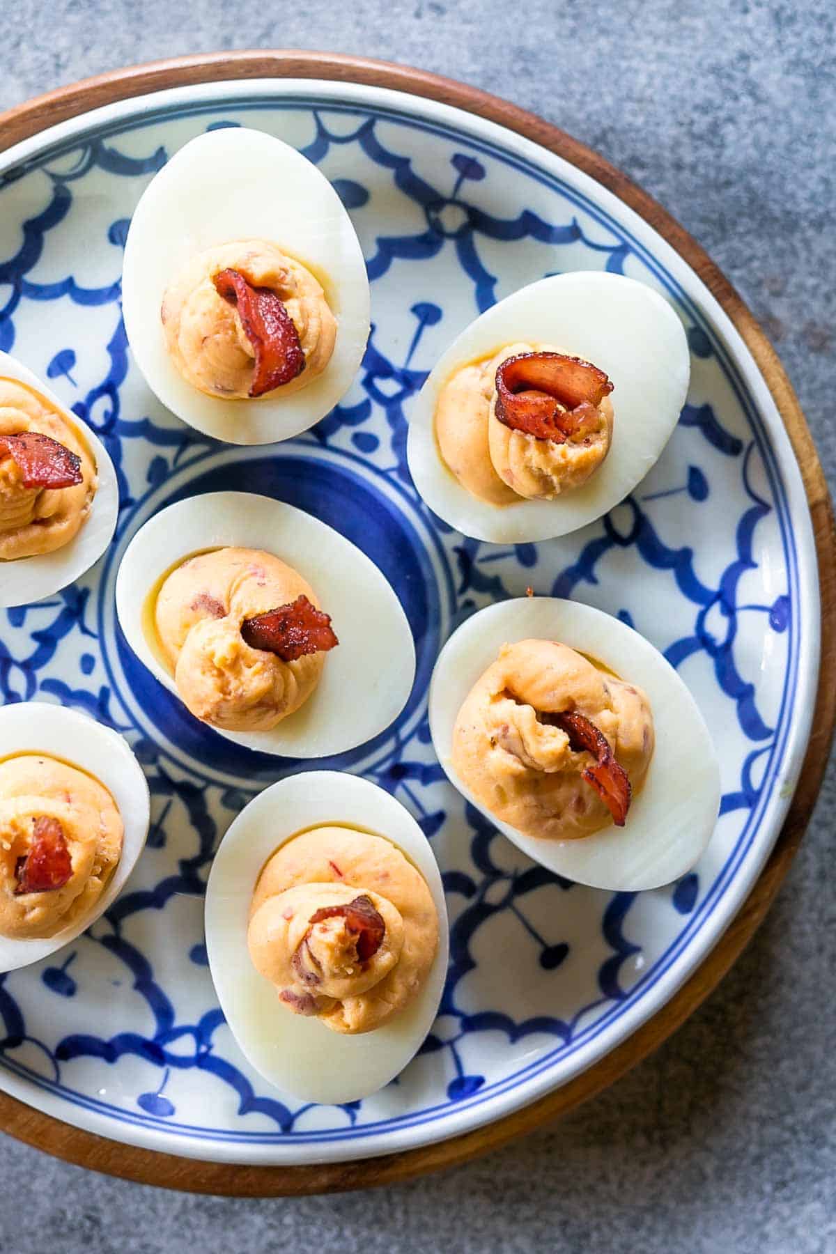 If you think regular deviled eggs are great, these spicy bacon deviled eggs are the bomb! They have a kick from the heat, and smokiness from the bacon and are really fast to put together. #eggs #brunch #easter #spring #recipes #appetizer #snack Spicy bacon deviled eggs on a blue and white plate topped with bacon