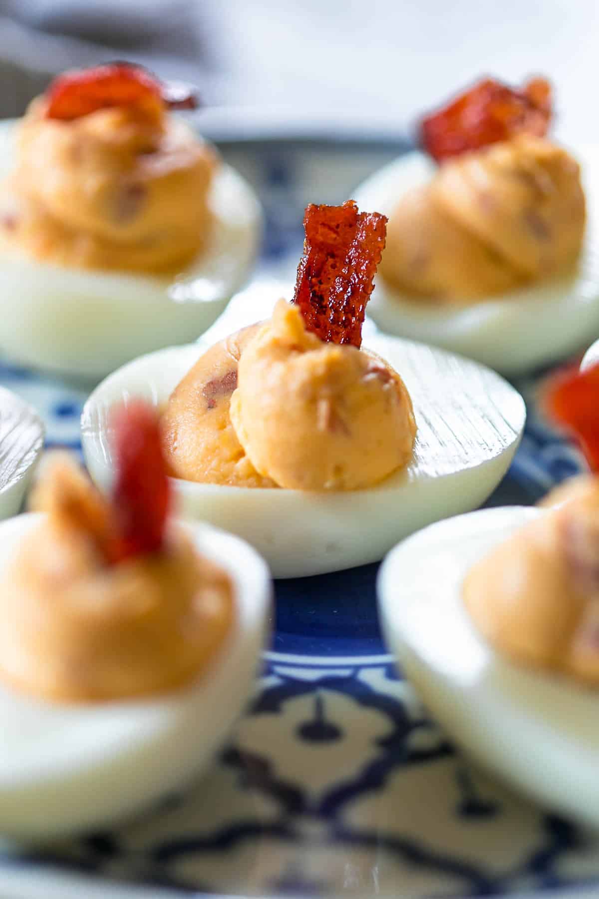 If you think regular deviled eggs are great, these spicy bacon deviled eggs are the bomb! They have a kick from the heat, and smokiness from the bacon and are really fast to put together. #eggs #brunch #easter #spring #recipes #appetizer #snack Close up of Spicy bacon deviled eggs on a blue and white plate topped with bacon