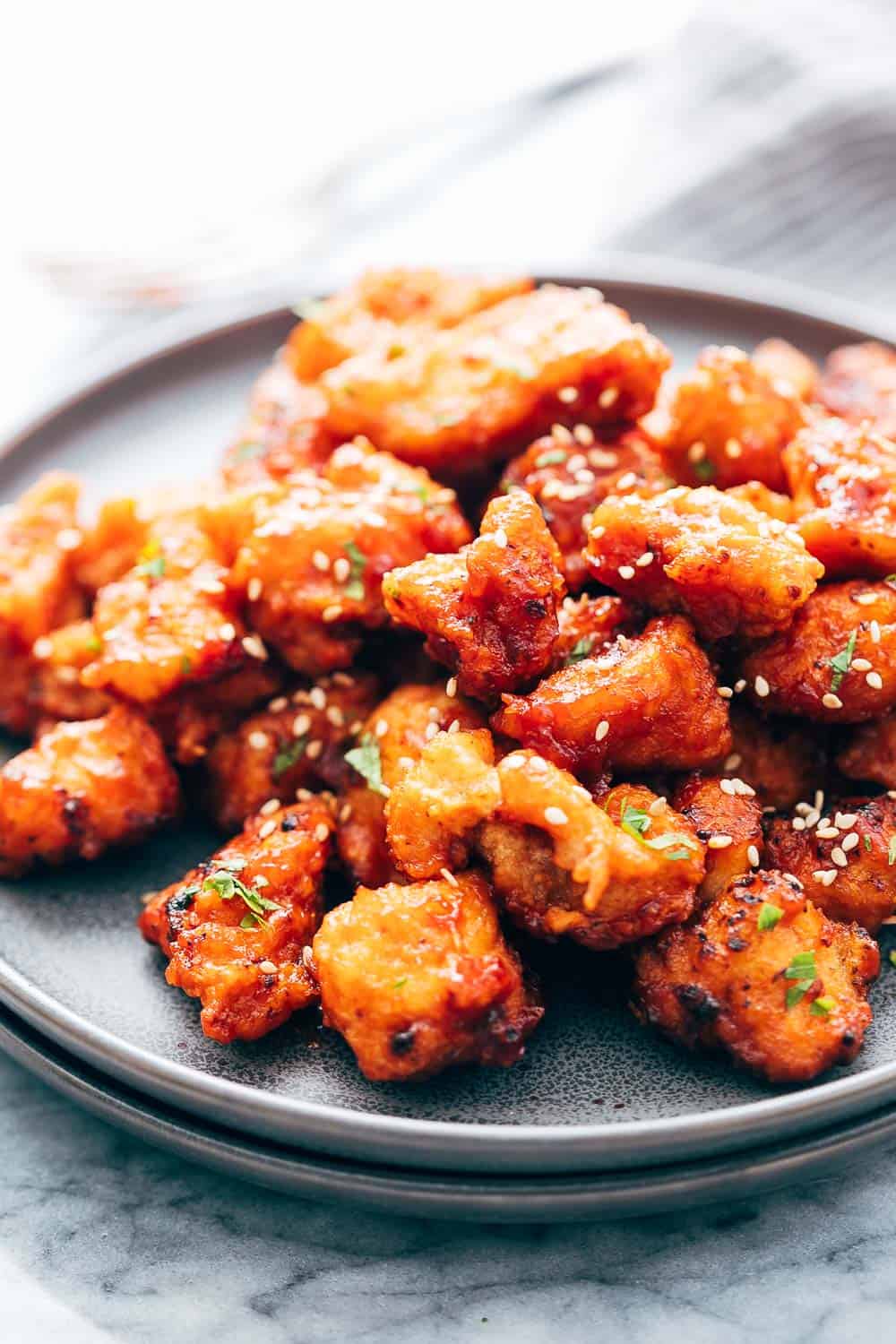 KOREAN POPCORN CHICKEN is sticky, spicy, tangy and crunchy. Inspired by the hyper-famous Korean street snack, Dakgangjeong, boneless chicken is deep fried and tossed in a sweet, sour and spicy sauce that makes every bite super addictive. #chicken #appetizer #party #snack #recipe Closeup of Korean Popcorn Chicken on a grey ceramic plate topped with sesame seeds and chopped parsley