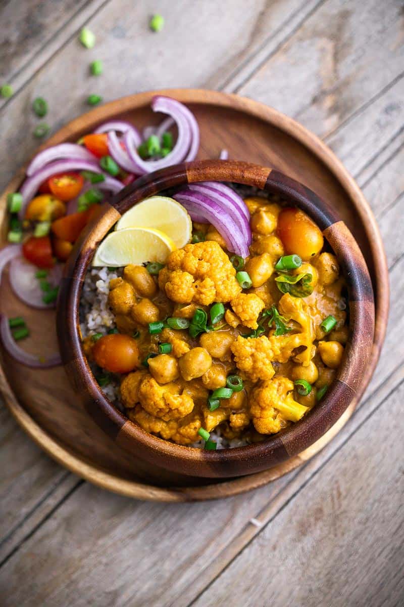 This is a classic coconut curry with tender cauliflower and soft chickpeas thats all made in one pot and has a simple mix of spices. That sauce is super addictive and you'll love how easy it is to make. It's vegan and gluten free, but you can easily add chicken, shrimp, lamb or beef if you prefer meat. #curry #sauce #coconut #cauliflower #dinner Cauliflower Chickpea Coconut Curry served in a brown bowl with onions and lime wedges