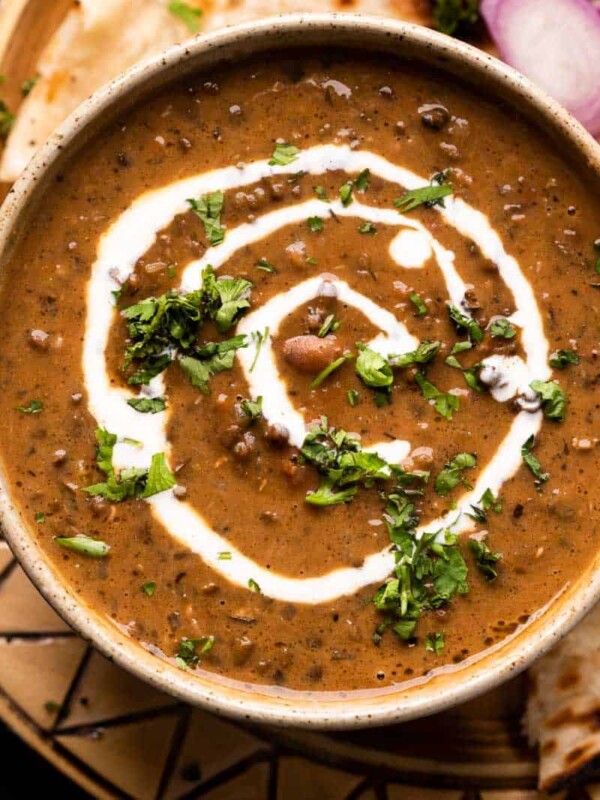 Dal Makhani served in a brown ceramic bowl with cream drizzled on top and naan and sliced onions on the side