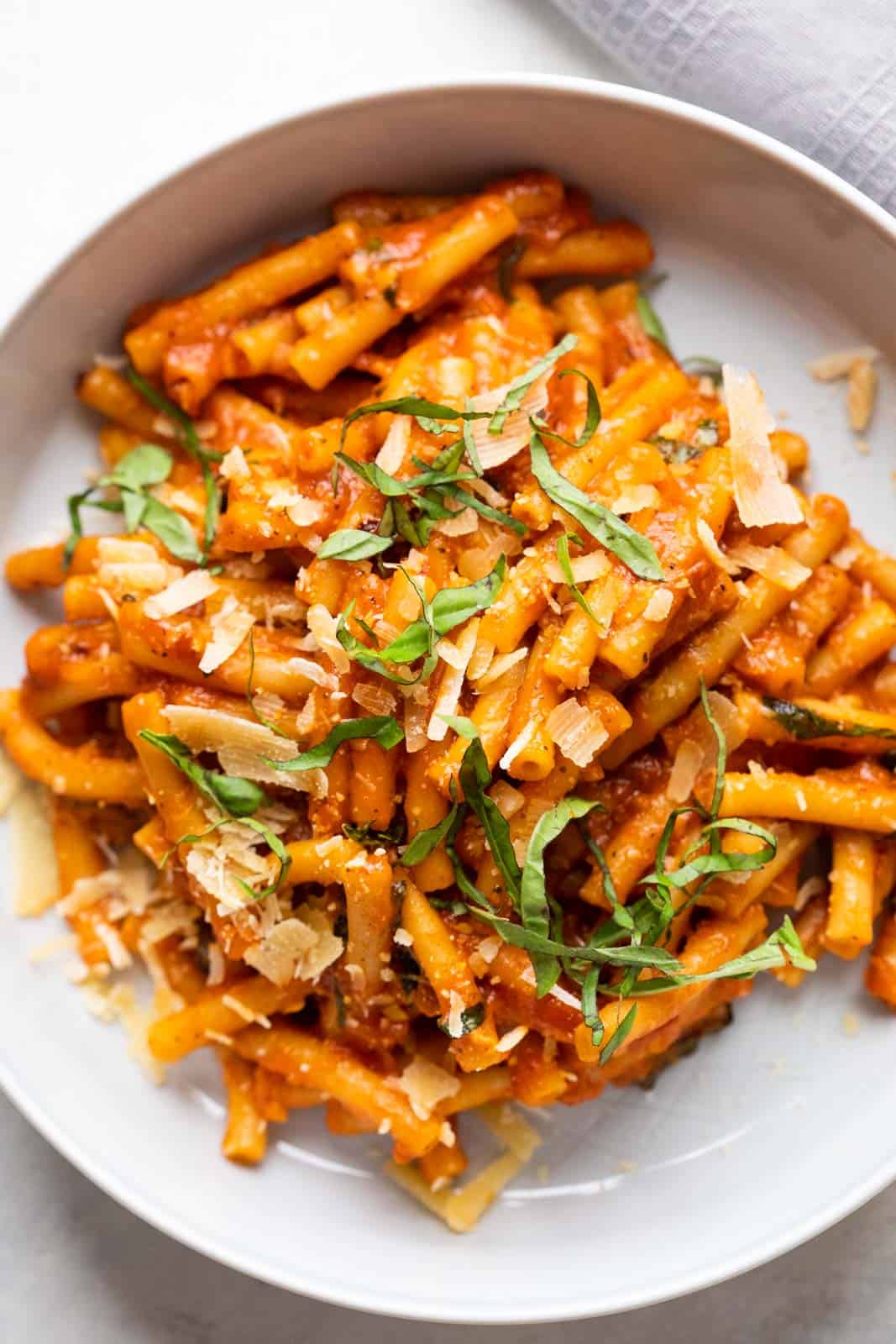Pasta in red sauce topped with shaved parmesan and basil served in a grey white bowl with a fork on the side