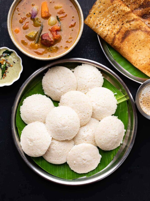 A picture of soft idlis stacked on top of each other on a banana leaf lined plate with dosas, chutney and sambar on the side