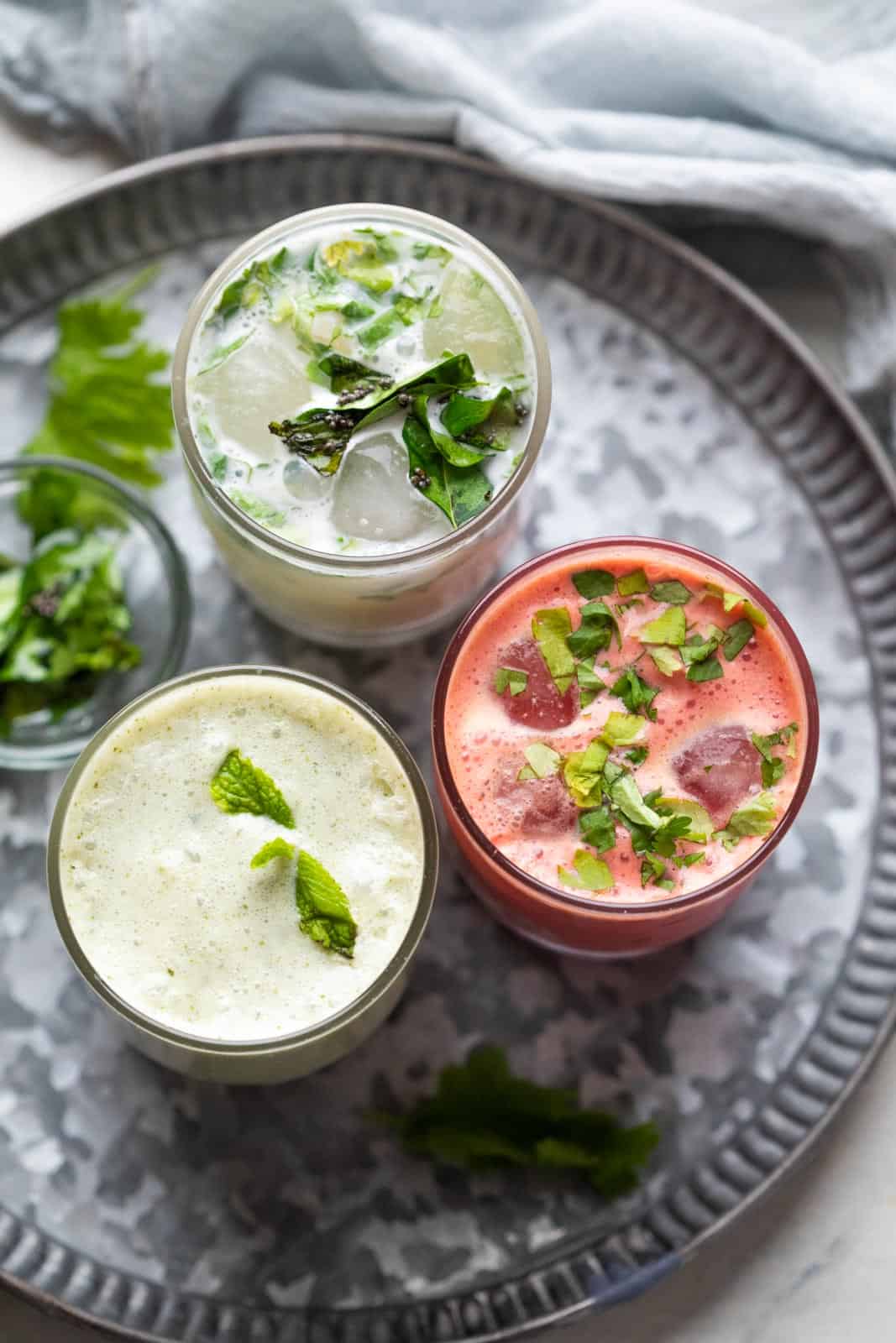 Three glasses of chaas served on a metal plate with curry leaves and coriander on the side