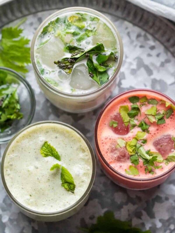 Three glasses of chaas served on a metal plate with curry leaves and coriander on the side