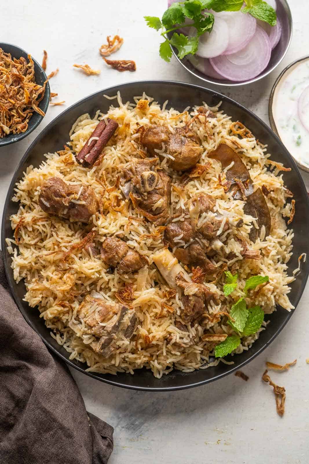 Mutton yakhni pulao served on a black plate with onions and raita on the side