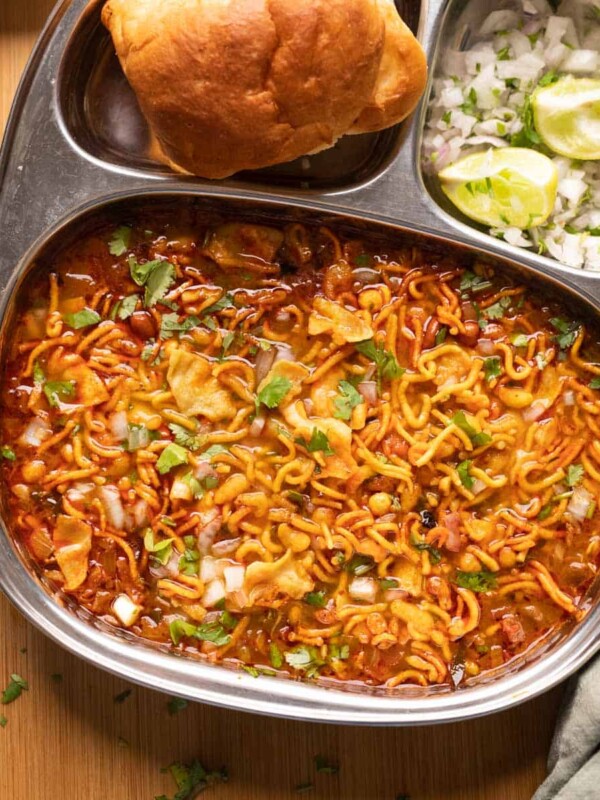 Picture of misal topped with farsaan served in a steel plate with pan, chopped onions and lime wedges on the side