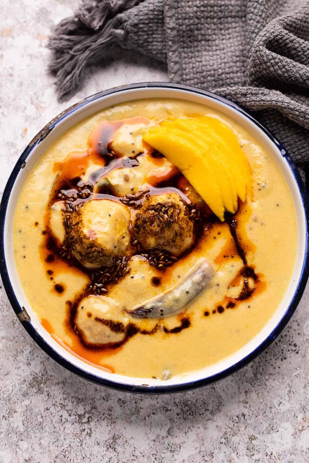 Mango Kadhi served in a blue and white enamel bowl with mango slices and tadka on top