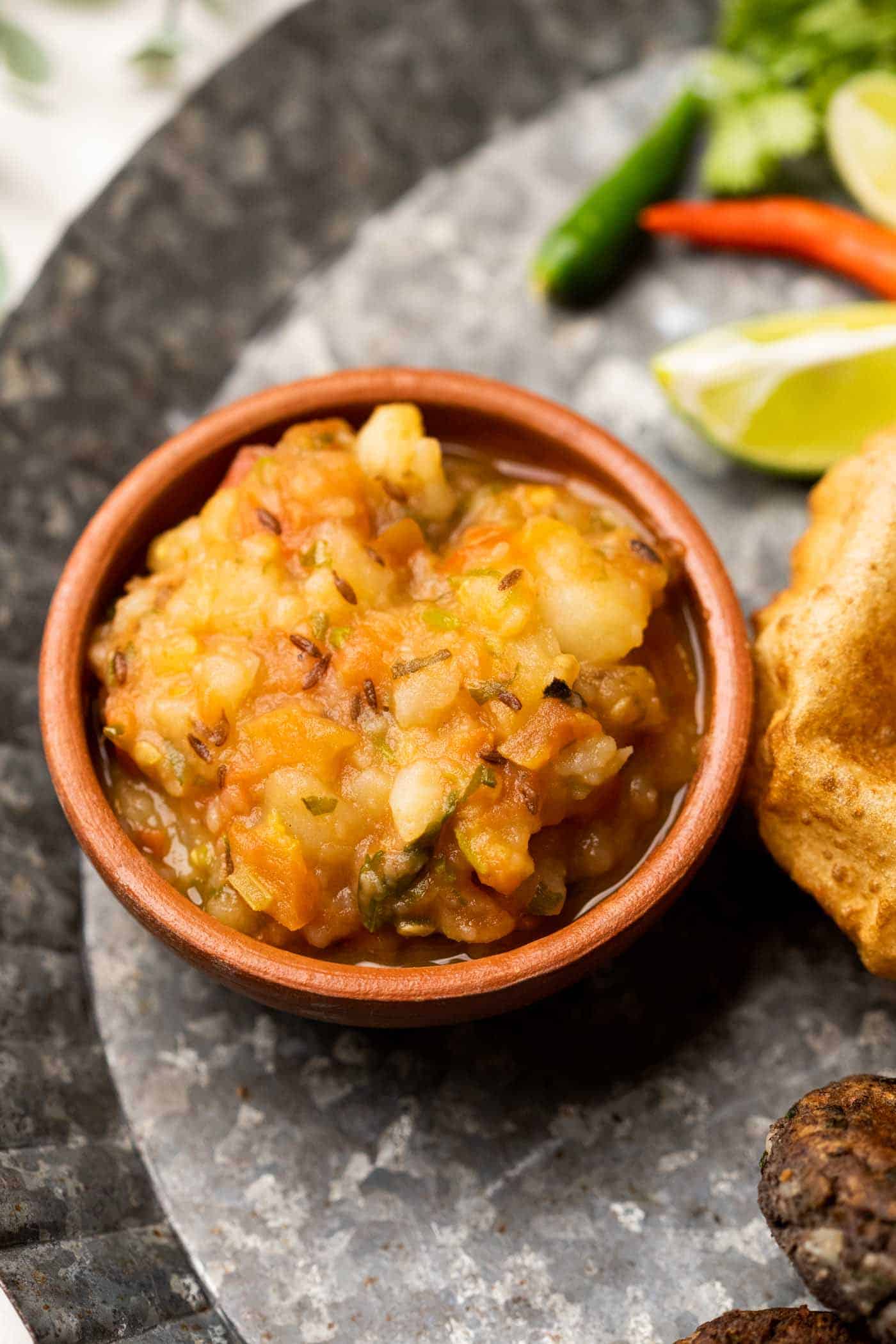Picture of potato curry served in a brown terracotta bowl with puri on the side