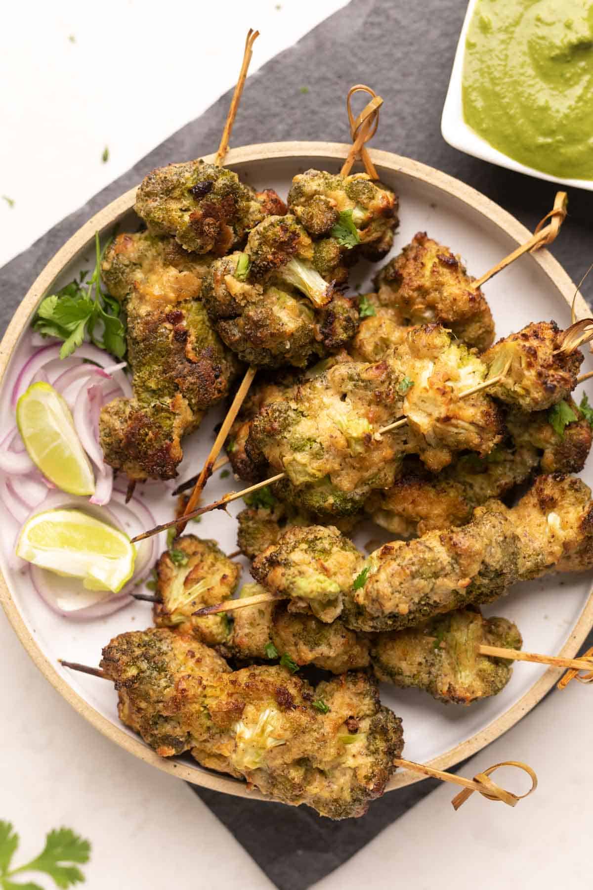 plate of malai broccoli on skewers with a side of sliced onion and green chutney