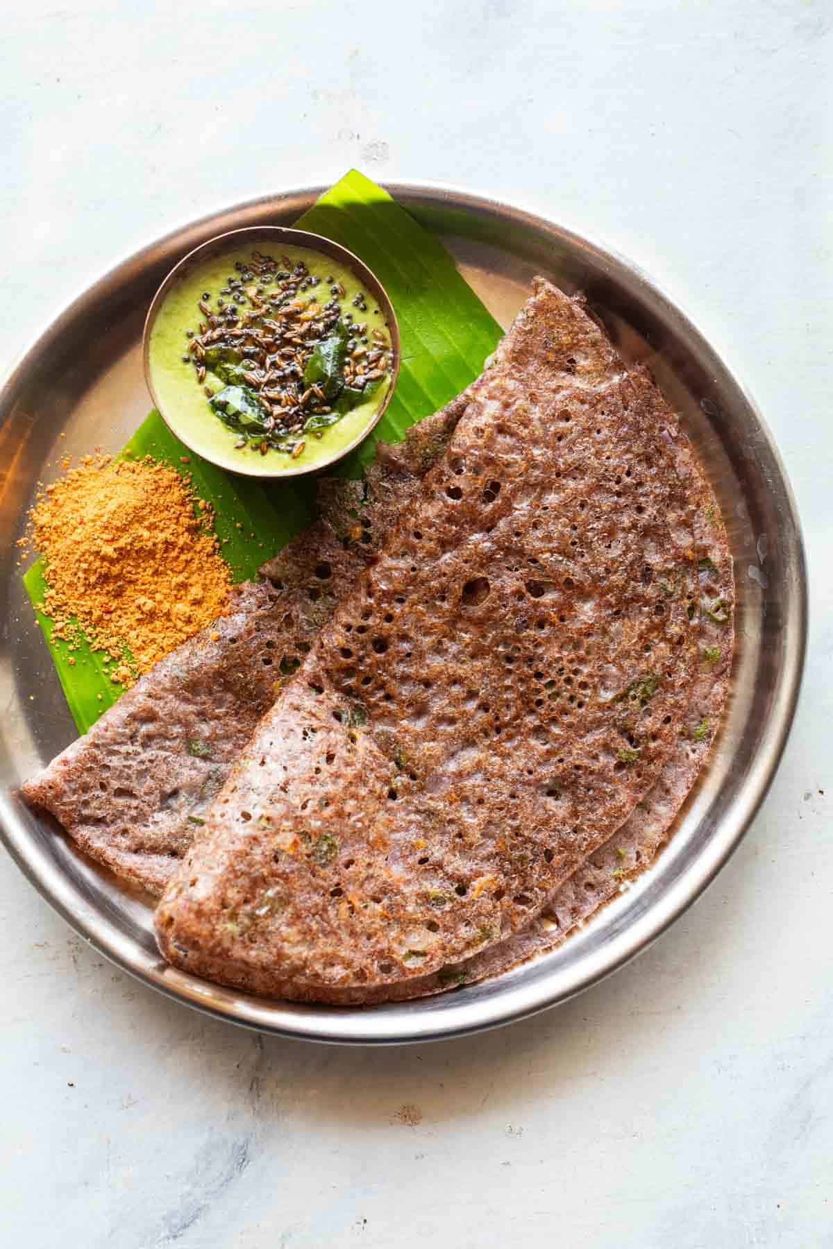 Ragi dosa served in a steel plate on a banana leaf with coconut chutney and podi