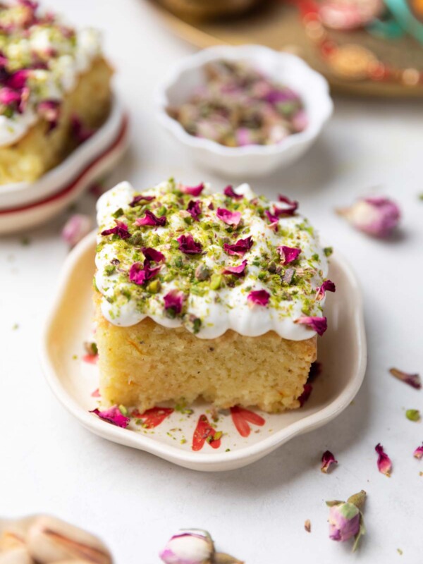 a slice of rasmalai cake garnished with cream, crushed nuts and rose petals on a white plate