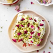 a slice of rasmalai cake garnished with cream, crushed nuts and rose petals on a white plate