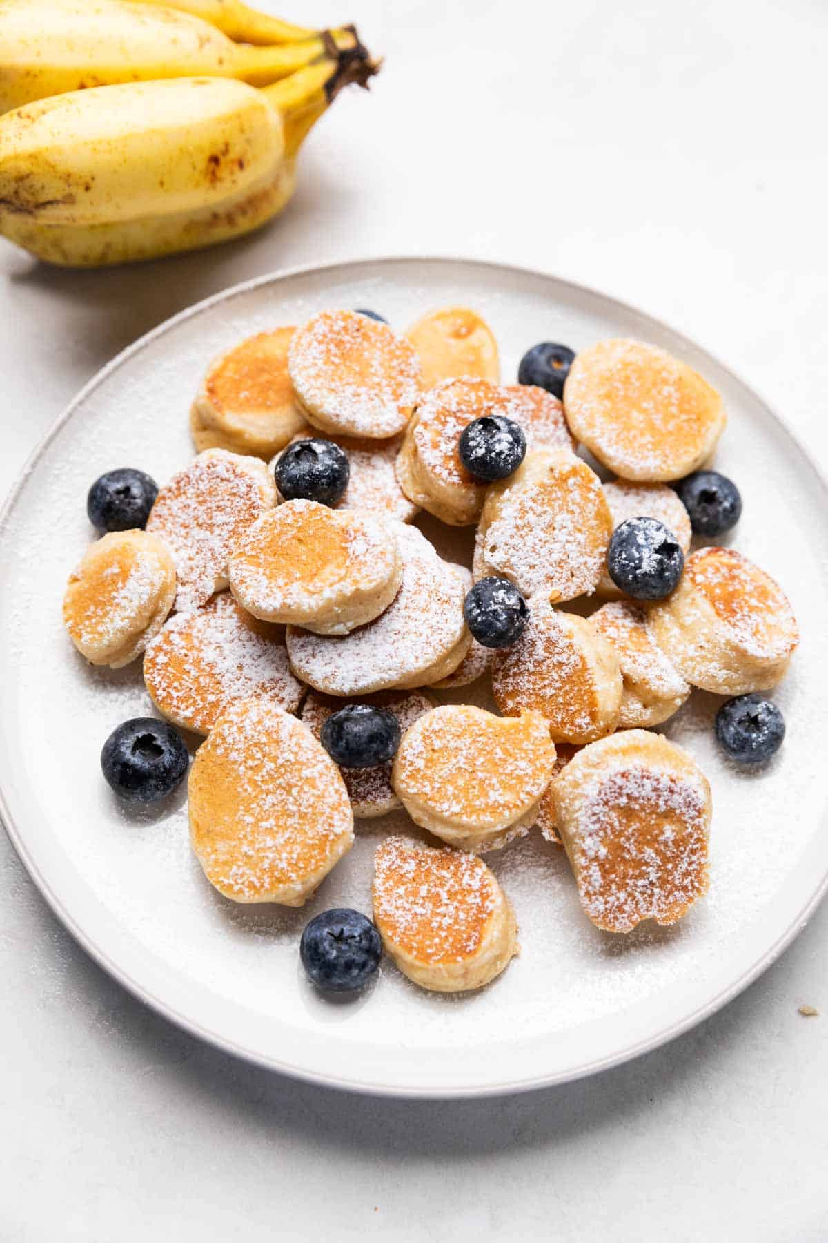 mini banana pancake bites garnished with powdered sugar & blueberries served on a white plate