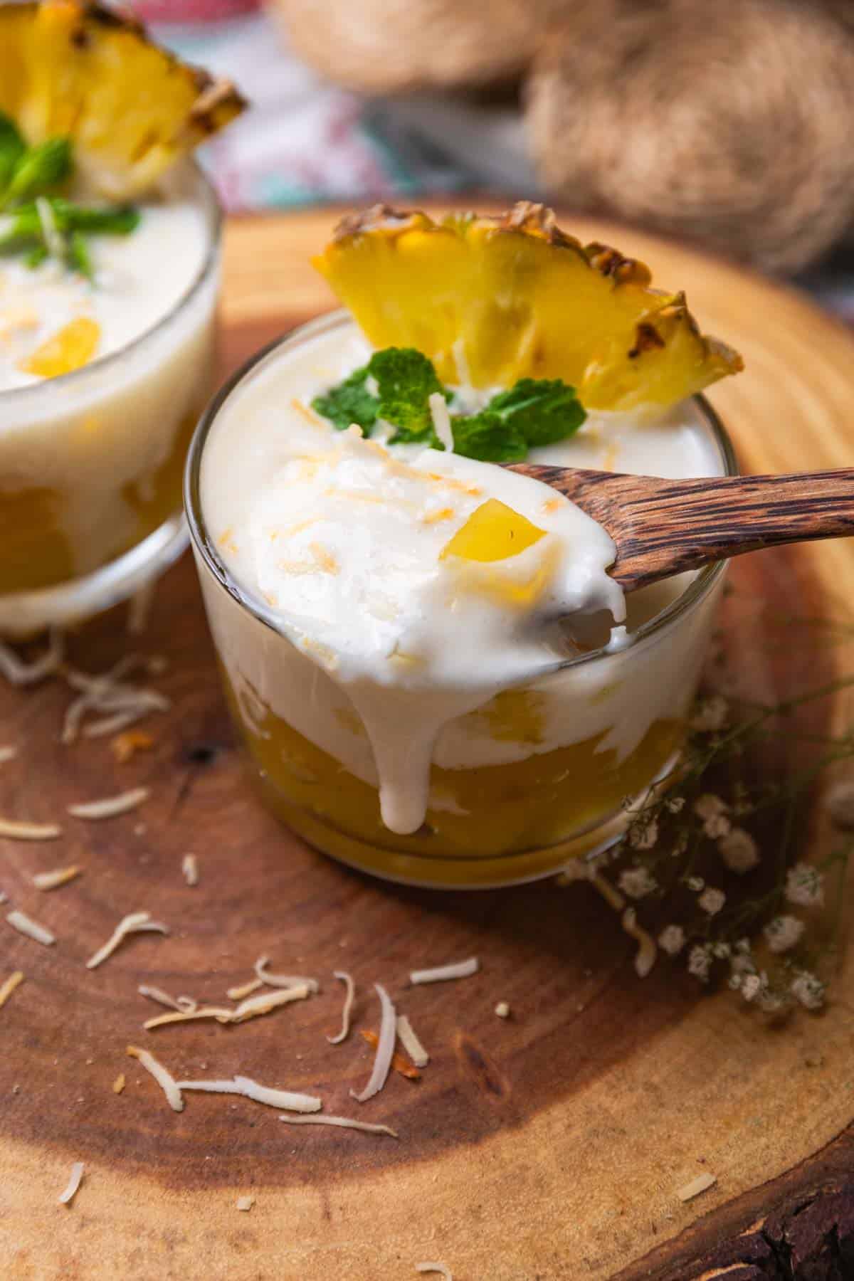 coconut pudding with pineapple jelly garnished with toasted coconut flakes, pineapple slice and mint leaves and served in shot glasses and a wooden spoon